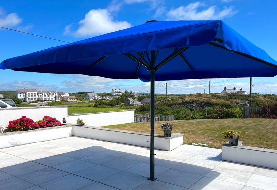 A blue parasol provides shade over a terrace with flowers and a rural landscape.